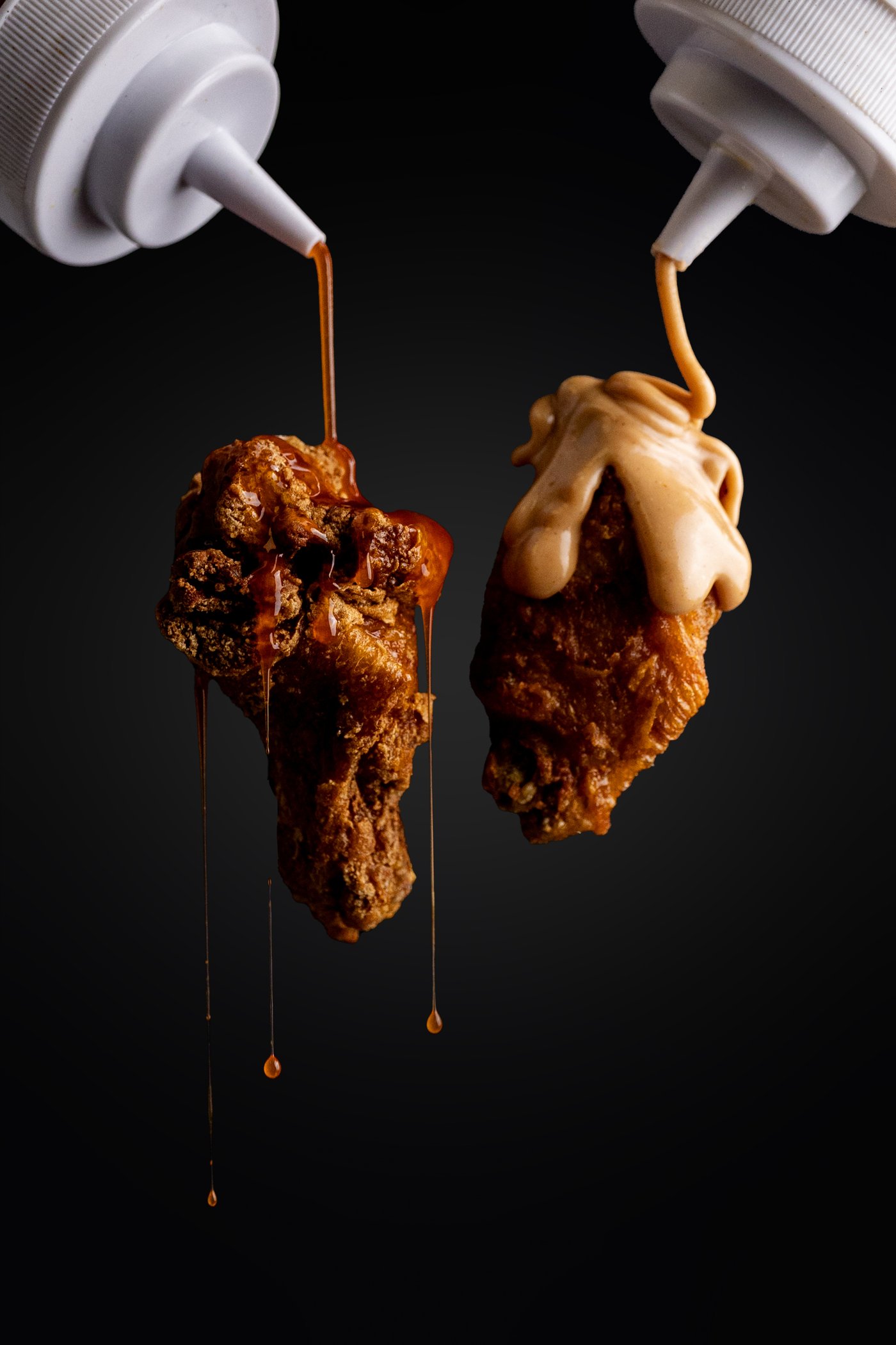 Two fried chicken wings mid-air, one drizzled with hot sauce and one with white sauce from squeeze bottles, shot by Wildcat