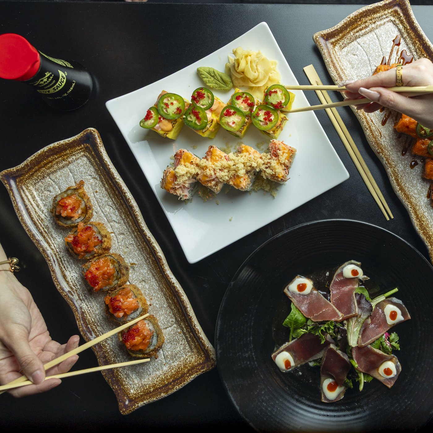 Overhead table of sushi rolls and tuna tataki with hands reaching in with chopsticks, shot for Sushi Lounge